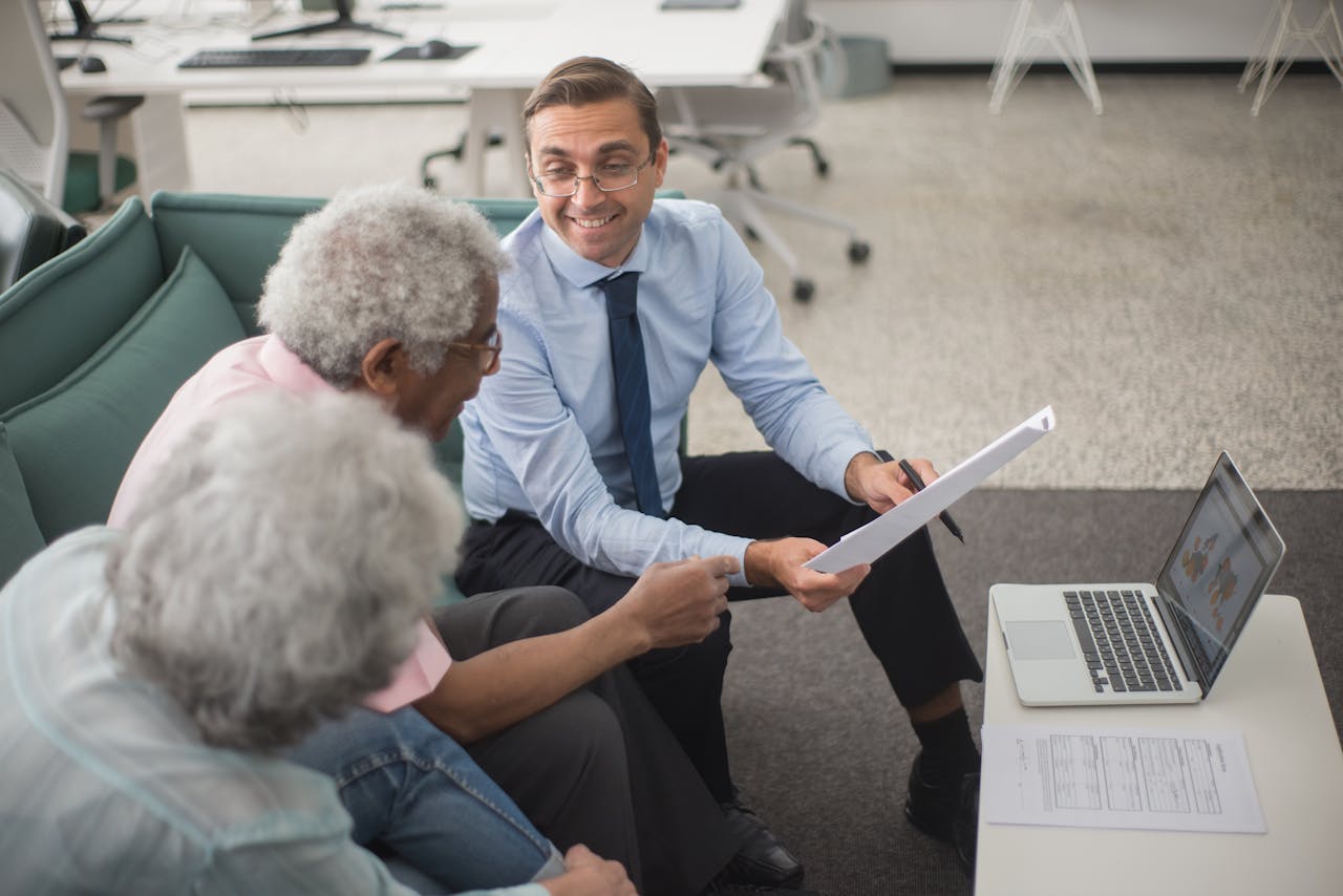 our-story Consultant discussing financial plans with senior clients in a modern office setting, using documents and a laptop.